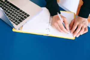 Une personne écrit dans un carnet près d’un ordinateur